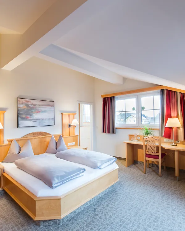 Helles Hotelzimmer mit Doppelbett, Schreibtisch, großem Fenster und warmen Holzmöbeln